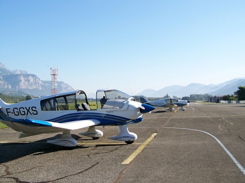 Two small, single-engine aircraft are parked on a tarmac near a mountain range. The planes have white and blue exteriors with visible registration numbers. The sky is clear, and the background features lush green fields and mountainous terrain.