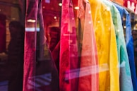 A display of brightly colored shirts hanging in a storefront. The shirts range in colors from red, pink, orange, yellow, green, to blue, creating a visually appealing gradient. The reflection of the street and passersby is visible in the glass.