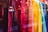 A display of brightly colored shirts hanging in a storefront. The shirts range in colors from red, pink, orange, yellow, green, to blue, creating a visually appealing gradient. The reflection of the street and passersby is visible in the glass.
