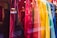 A display of brightly colored shirts hanging in a storefront. The shirts range in colors from red, pink, orange, yellow, green, to blue, creating a visually appealing gradient. The reflection of the street and passersby is visible in the glass.