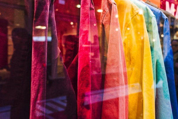 A display of brightly colored shirts hanging in a storefront. The shirts range in colors from red, pink, orange, yellow, green, to blue, creating a visually appealing gradient. The reflection of the street and passersby is visible in the glass.