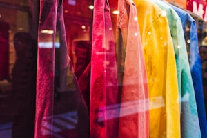 A display of brightly colored shirts hanging in a storefront. The shirts range in colors from red, pink, orange, yellow, green, to blue, creating a visually appealing gradient. The reflection of the street and passersby is visible in the glass.