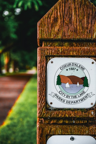A wooden signpost featuring an emblem with the text 'Coeur d'Alene 1887 City by the Lake Parks Department'. The emblem depicts a lake scene with sailboats and trees against a mountainous background. The surroundings include lush green foliage and a pathway.