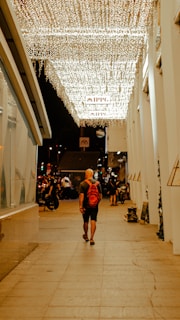 A person with a red backpack walks underneath a canopy of glowing string lights. The scene is set in an urban environment with a mixture of modern architecture and street activity. The surrounding area is subtly illuminated by the lights, creating a warm ambiance.