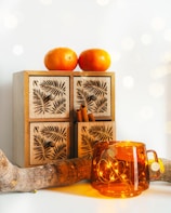 A wooden box with intricate leaf carvings has two oranges placed on top. Cinnamon sticks are visible from one of the box drawers, while a piece of driftwood is positioned on a flat surface. An amber-colored glass cup containing warm, glowing string lights adds a cozy ambiance.