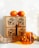 A wooden box with intricate leaf carvings has two oranges placed on top. Cinnamon sticks are visible from one of the box drawers, while a piece of driftwood is positioned on a flat surface. An amber-colored glass cup containing warm, glowing string lights adds a cozy ambiance.