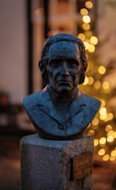 A bronze bust of a person is placed on a stone pedestal against a blurred background of warm, glowing lights. The bust depicts a man in a suit, with detailed facial features and short hair.