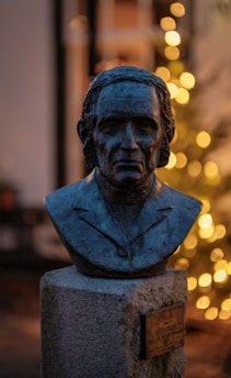 A bronze bust of a person is placed on a stone pedestal against a blurred background of warm, glowing lights. The bust depicts a man in a suit, with detailed facial features and short hair.