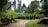 A lush garden with various potted plants and bonsai trees arranged on stands. The garden features a stone pathway leading through the greenery. Tall trees and dense foliage surround the area, with some skyscrapers visible in the background sky. Sunlight casts dappled shadows across the scene.