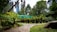 A lush garden with various potted plants and bonsai trees arranged on stands. The garden features a stone pathway leading through the greenery. Tall trees and dense foliage surround the area, with some skyscrapers visible in the background sky. Sunlight casts dappled shadows across the scene.