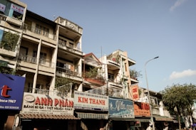 A row of urban buildings line the street, with multiple levels and balconies. Storefronts on the ground floor display various signs in Vietnamese. Some of the buildings have plants and laundry on the balconies. The streetlamp and trees indicate a well-populated area. The sky is clear with a few clouds.