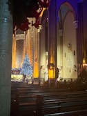 A dimly lit church interior with tall, ornate columns and stained glass windows. Decorations include a brightly lit Christmas tree adorned with blue and white lights, surrounded by festive garlands and wreaths. Empty pews lead to the altar, which is bathed in a warm orange glow.