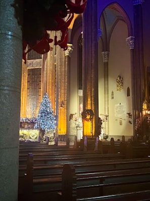 A dimly lit church interior with tall, ornate columns and stained glass windows. Decorations include a brightly lit Christmas tree adorned with blue and white lights, surrounded by festive garlands and wreaths. Empty pews lead to the altar, which is bathed in a warm orange glow.