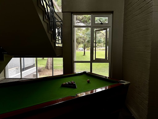 A pool table is positioned under a staircase in a dimly lit room. Natural light illuminates the area through a large window with a view of a grassy outdoor space and trees. Pool balls are arranged on the green felt surface of the table.