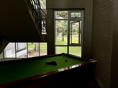 A pool table is positioned under a staircase in a dimly lit room. Natural light illuminates the area through a large window with a view of a grassy outdoor space and trees. Pool balls are arranged on the green felt surface of the table.