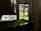 A pool table is positioned under a staircase in a dimly lit room. Natural light illuminates the area through a large window with a view of a grassy outdoor space and trees. Pool balls are arranged on the green felt surface of the table.