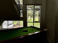 A pool table is positioned under a staircase in a dimly lit room. Natural light illuminates the area through a large window with a view of a grassy outdoor space and trees. Pool balls are arranged on the green felt surface of the table.