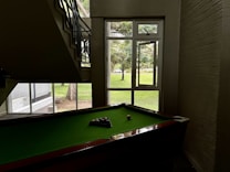 A pool table is positioned under a staircase in a dimly lit room. Natural light illuminates the area through a large window with a view of a grassy outdoor space and trees. Pool balls are arranged on the green felt surface of the table.