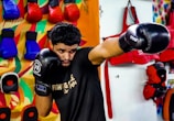 A man wearing black boxing gloves is practicing in a brightly colored gym. He is throwing a punch with his right hand while his left hand guards his face. In the background, there are variously colored training pads and gloves hanging on the wall. The setting includes a vibrant, multi-colored mural and several pieces of training equipment organized on the wall.