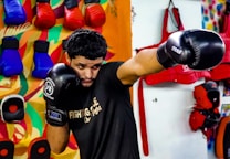 A man wearing black boxing gloves is practicing in a brightly colored gym. He is throwing a punch with his right hand while his left hand guards his face. In the background, there are variously colored training pads and gloves hanging on the wall. The setting includes a vibrant, multi-colored mural and several pieces of training equipment organized on the wall.