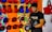 A person wearing a black t-shirt is in a room with numerous colorful boxing gloves hanging on the wall. The wall has a vibrant and artistic background with abstract patterns. The person appears to be handling black and red boxing equipment.