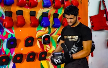 A person wearing a black t-shirt is in a room with numerous colorful boxing gloves hanging on the wall. The wall has a vibrant and artistic background with abstract patterns. The person appears to be handling black and red boxing equipment.