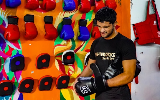 A person wearing a black t-shirt is in a room with numerous colorful boxing gloves hanging on the wall. The wall has a vibrant and artistic background with abstract patterns. The person appears to be handling black and red boxing equipment.