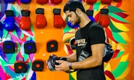 A man standing in front of a colorful wall holds a pair of black boxing gloves. The wall features vibrant painted patterns and is lined with red and blue boxing gloves.