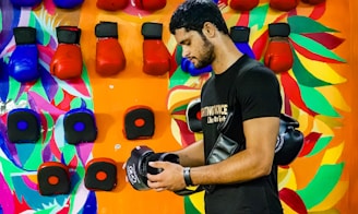 A man standing in front of a colorful wall holds a pair of black boxing gloves. The wall features vibrant painted patterns and is lined with red and blue boxing gloves.