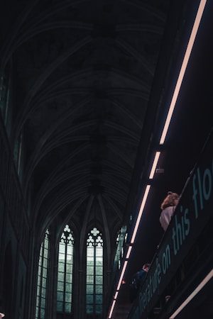 A dark, gothic-style interior with large, ornate stained glass windows. The architecture features arched ceilings and detailed stonework. A balcony or upper floor is visible with a person gazing down. The lighting is moody, with a contrast between the dim atmosphere and the bright modern lights.