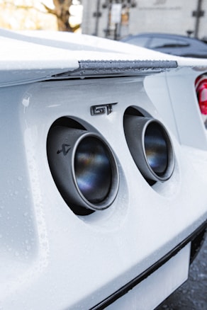 Close-up of a white sports car's rear exhaust system, featuring dual exhaust pipes with a metallic sheen and water droplets on the car's surface. The 'GT' emblem is visible near the pipes, and part of another car is seen in the blurred background.