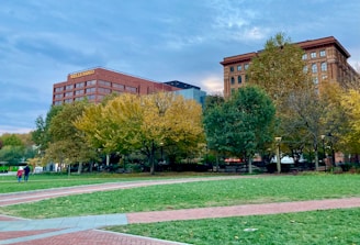 A cityscape featuring two prominent buildings with one labeled with a bank's name. The scene includes a park in the foreground with lush green grass, autumn-colored trees, and paved walkways. A few people are strolling along the pathways, contributing to a peaceful urban environment.