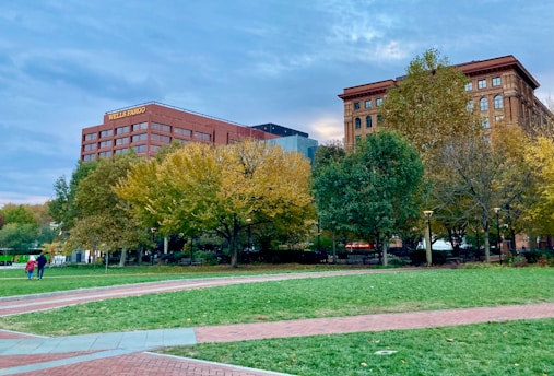 A cityscape featuring two prominent buildings with one labeled with a bank's name. The scene includes a park in the foreground with lush green grass, autumn-colored trees, and paved walkways. A few people are strolling along the pathways, contributing to a peaceful urban environment.