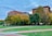 A cityscape featuring two prominent buildings with one labeled with a bank's name. The scene includes a park in the foreground with lush green grass, autumn-colored trees, and paved walkways. A few people are strolling along the pathways, contributing to a peaceful urban environment.