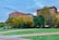 A cityscape featuring two prominent buildings with one labeled with a bank's name. The scene includes a park in the foreground with lush green grass, autumn-colored trees, and paved walkways. A few people are strolling along the pathways, contributing to a peaceful urban environment.