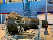 A historic spacecraft capsule displayed indoors, featuring a rounded, metallic body with riveted panels and a large window. The words 'UNITED STATES' are prominently painted on the side. The capsule is mounted on a supportive frame for exhibition, and the surrounding area includes a blue wall and some structural elements.