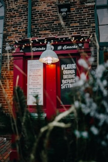 A rustic brick building features a bright red exterior window with the words 'Drinks and Snacks' on top. A blackboard sign reads 'Please order inside before sitting'. The window is lit with a soft yellow light under a vintage-style lamp. Holiday decorations, including small white lights, add a festive touch.