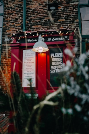 A rustic brick building features a bright red exterior window with the words 'Drinks and Snacks' on top. A blackboard sign reads 'Please order inside before sitting'. The window is lit with a soft yellow light under a vintage-style lamp. Holiday decorations, including small white lights, add a festive touch.