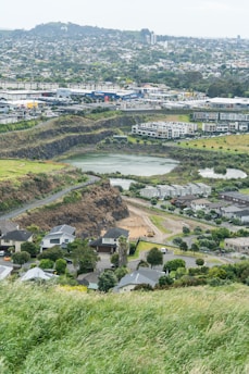 A sprawling suburban landscape with a mixture of residential houses in the foreground and commercial buildings in the distance. A grassy hill descends into a neighborhood with modern houses, surrounded by trees and roads. Further back, an industrial area with large warehouse buildings and parking areas can be seen. In the background, a densely forested hill overlooks the town.