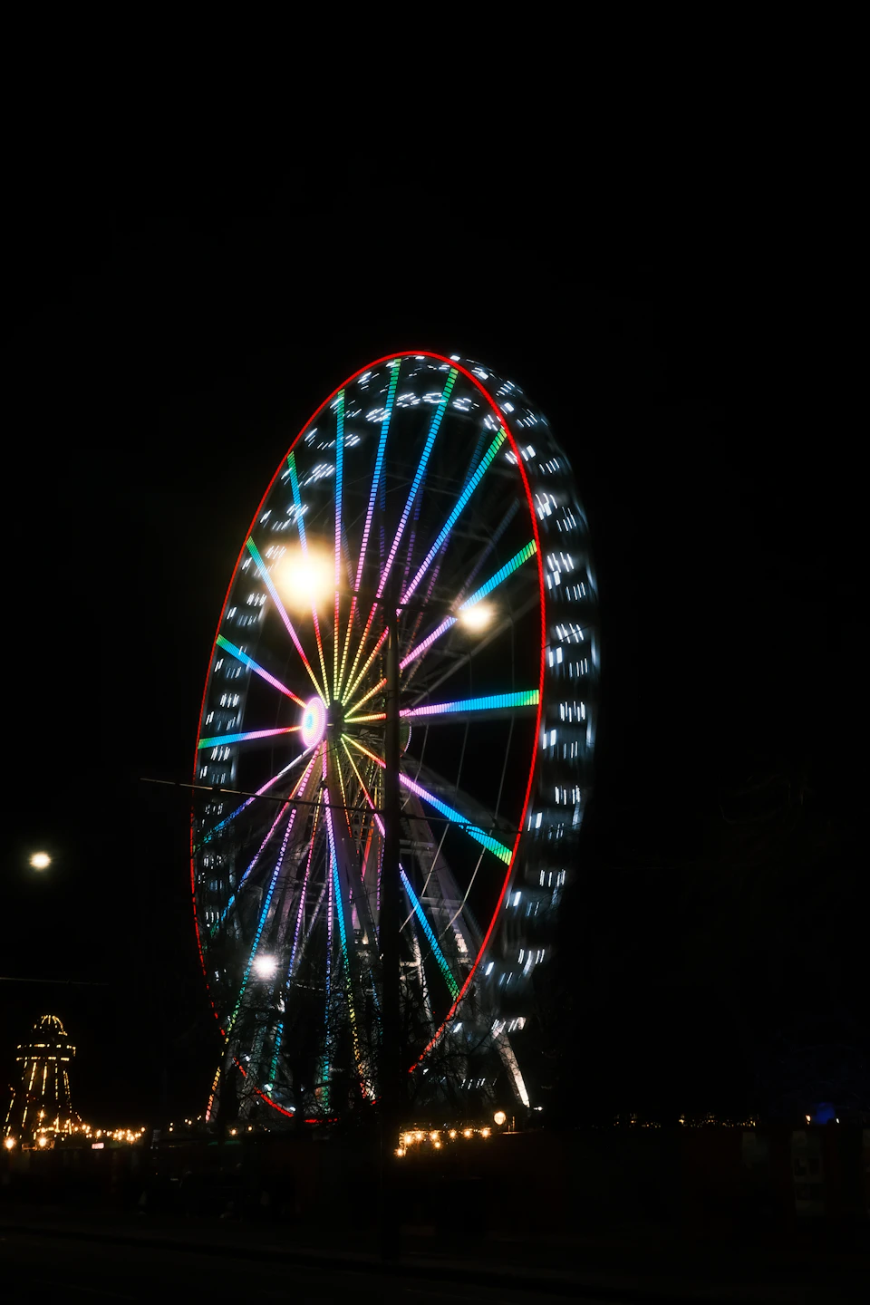 The image depicts a large, brightly lit Ferris wheel at night. The Ferris wheel is illuminated with vibrant colors and stands out against the dark sky. In the background, there are other lights that add to the festive atmosphere. A few people can be seen in the distance, possibly enjoying the view or taking photos of the impressive structure.