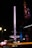 A tall, illuminated amusement ride stands against the night sky, surrounded by colorful lights that create a vibrant, dynamic scene. Adjacent to it is a dimly lit Gothic-style structure with intricate details, highlighted by blue and purple lighting.