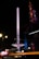 A tall, illuminated amusement ride stands against the night sky, surrounded by colorful lights that create a vibrant, dynamic scene. Adjacent to it is a dimly lit Gothic-style structure with intricate details, highlighted by blue and purple lighting.