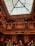 An intricately detailed wooden balcony showcases elaborate carving and architectural craftsmanship, set against a backdrop of a glass ceiling. The warm lighting accentuates the ornate patterns and rich textures of the woodwork.