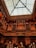 An intricately detailed wooden balcony showcases elaborate carving and architectural craftsmanship, set against a backdrop of a glass ceiling. The warm lighting accentuates the ornate patterns and rich textures of the woodwork.