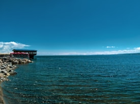 A serene lakeside view with a wooden cabin-like structure perched on stilts over the water, surrounded by rocky shores. The sky is clear and blue, reflecting its hue onto the expansive water surface.