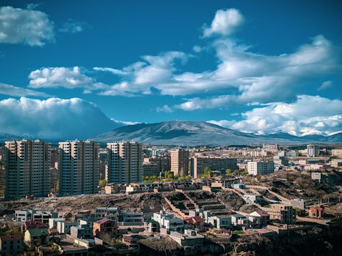 A cityscape featuring high-rise apartment buildings in the foreground with a sprawling urban area. In the background, a range of mountains under a partly cloudy sky suggests a natural setting surrounding the city.