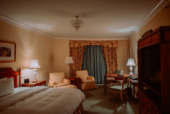 A well-furnished bedroom with a plush bed, a large chandelier hanging from the ceiling, and elegant decor. Two armchairs and a round table are placed in front of a window with heavy floral curtains. There is a wooden desk and chair near a lamp, and a large wooden cabinet on the opposite side includes a television. The lighting is soft and warm, creating a cozy atmosphere.