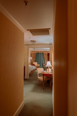 A narrow hallway leads into a warmly lit hotel room with classic decor. The room features a bed with white linens and a wooden headboard, a small wooden desk with a chair, and a lamp emitting a soft glow. Curtains with intricate patterns add a touch of elegance. The carpet is green, complementing the neutral tones of the room.