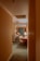 A narrow hallway leads into a warmly lit hotel room with classic decor. The room features a bed with white linens and a wooden headboard, a small wooden desk with a chair, and a lamp emitting a soft glow. Curtains with intricate patterns add a touch of elegance. The carpet is green, complementing the neutral tones of the room.