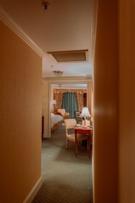 A narrow hallway leads into a warmly lit hotel room with classic decor. The room features a bed with white linens and a wooden headboard, a small wooden desk with a chair, and a lamp emitting a soft glow. Curtains with intricate patterns add a touch of elegance. The carpet is green, complementing the neutral tones of the room.
